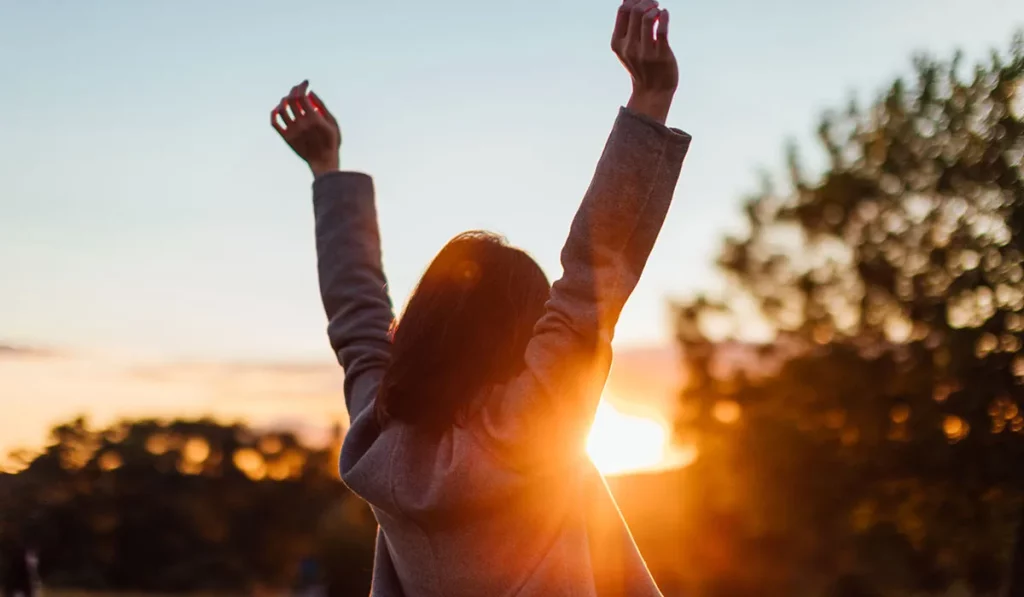 Person enjoying sunset with arms raised in the air, basking in warm sunlight outdoors.